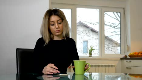 Woman-sitting-in-the-kitchen-and-scrolling-the-phone-while-drinking-coffee