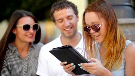 Group-of-Students-with-Tablet-Hanging-out.-Girl-in-Light-Blue-Dress-is-Reading-Something,-Her-Female-Friend-and-Boyfriend-Listen-Sitting-Outdoors-During-Warm-Sunny-Day-in-the-City