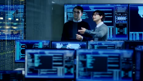 In-the-System-Control-Room-Project-Manage-and-IT-Engineer-Have-Discussion,-they're-surrounded-by-Multiple-Monitors-with-Graphics.-They-Work-in-a-Data-Center-on-Data-Mining,-AI-and-Neural-Networking.