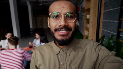 Close-up-of-afro-americano-hipster-hombre-en-vasos-con-smartphone-y-tener-video-llamada-onlive-mientras-sus-compañeros-trabajan-en-la-oficina-moderna