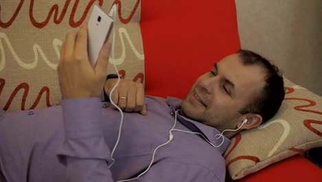 Young-Man-having-video-chat-holding-smartphone-webcam-chatting-to-girlfriend-lying-on-sofa-relaxing-at-home.