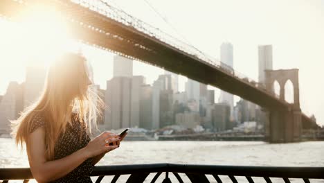 Hermosa-chica-con-el-pelo-largo-en-gafas-de-sol-usando-la-aplicación-para-smartphone-en-el-muelle-del-río-al-atardecer-cerca-de-puente-de-Brooklyn-Nueva-York-4K