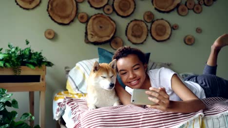 Fröhliches-Mädchen-nimmt-Selfie-mit-Haustier-posiert-mit-schöner-Hund-auf-Bett,-Spaß-haben-und-lachen.-Modernes-Apartment-mit-schönen-Design-und-Möbel-ist-sichtbar.