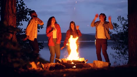 Excited-young-men-and-women-hipsters-are-dancing-around-bright-campfire-having-outdoor-party-in-forest-having-fun.-People,-nature-and-friendship-concept.