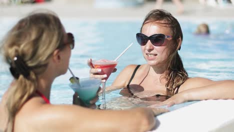 Zwei-Frauen-in-Sonnenbrille-sprechen-im-Pool-und-einen-Cocktail-zu-trinken.