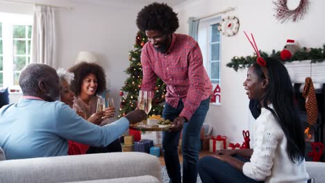 Hombre-que-sirve-champán-y-aperitivos,-como-los-miembros-adultos-de-la-familia-celebran-la-Navidad-en-casa