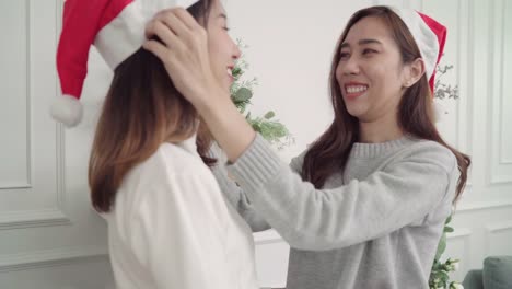 Pareja-de-lesbianas-asiáticas-dar-y-usar-sombrero-de-la-Navidad-uno-al-otro-en-su-sala-de-estar-en-el-país-en-el-Festival-de-Navidad.-Las-mujeres-del-estilo-de-vida-lgbt-felizes-celebran-Navidad-y-año-nuevo-concepto.