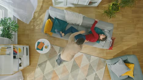 Young-Beautiful-Couple-is-Fooling-and-Partying-Around.-Running-Across-the-Room-and-Lying-Down-on-Sofa.-Cozy-Living-Room-with-Modern-Interior-with-Plants,-Table-and-Wooden-Floor.-Top-View.