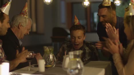 Happy-Smiling-Boy-Blowing-Candles-out-on-her-Birthday-Cake.-The-Boy-Surrounded-by-His-Family-and-Friends