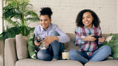 Two-mixed-race-curly-girl-friends-sitting-on-couch-play-console-computer-games-with-gamepad-and-have-fun-at-home