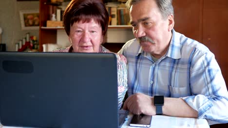 Una-pareja-de-ancianos-está-sentado-en-casa-en-la-computadora-portátil.-Una-mujer-lee-noticias,-un-hombre-con-un-bigote-se-sienta-junto-a-él-y-las-conversaciones