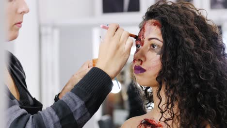 Make-up-artist-make-the-girl-halloween-make-upin-studio.Halloween-face-art.Woman-applies-on-professional-greasepaint-on-the-face-of-spanish-girl.War-paint-with-blood,-scars-and-wounds.-4k