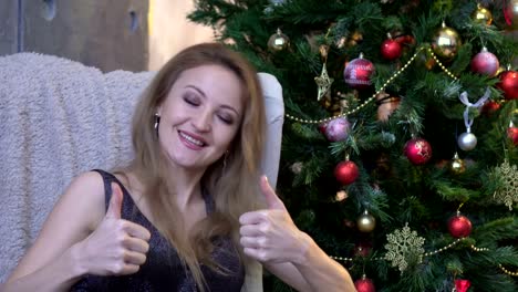 Joyful-woman-giving-two-thumbs-up-and-sitting-on-christmas-tree-background