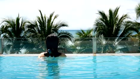 4K.-Asian-woman-springs-up-from-under-the-pool,-swimming-and-relax-at-the-pool-with-palm-tree-tropical-beach-at-background.-summer-recreation-at-tropical-beach-concept