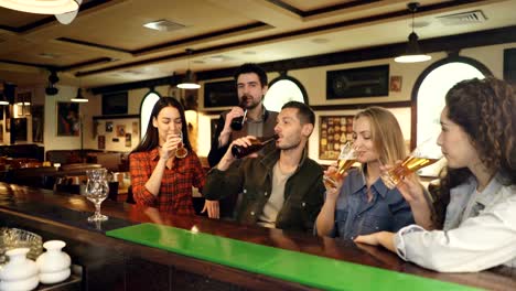 Young-people-are-celebrating-something-over-beer-in-bar.-They-are-proposing-toast,-clanging-glasses-and-bottles,-chatting-and-laughing.-Celebration-happiness-concept.