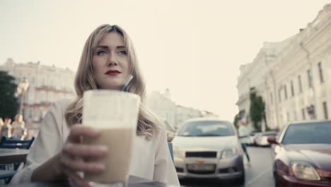 young-caucasian-blonde-woman,-curly-hair,-with-sexy-red-lips-drinking-an-alcoholic-cocktail-through-a-black-straw-from-a-large-bar-glass-in-a-restaurant-on-a-city-street.-Lonely-waiting-for-a-date