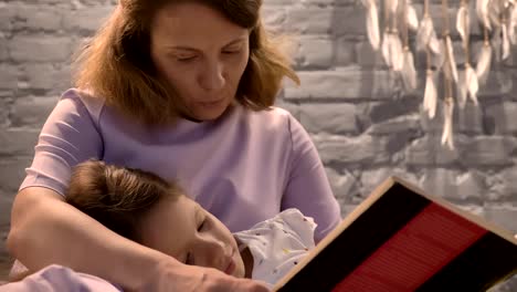 Mother-reading-book-to-little-daughter,-kid-lying-on-parents-laps-and-listening-tail,-family-concept-at-home,-indoors