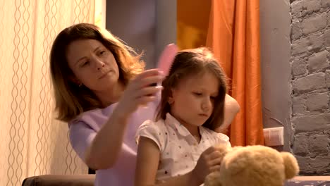 Mother-brushing-hair-of-her-little-daughter-in-modern-living-room,-kid-playing-with-teddy,-family-concept-indoors