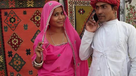 Tilt-up-to-Indian-couple-in-traditional-clothes-busy-on-a-phone-call-with-traditional-carpets-hanging-in-the-backdrop