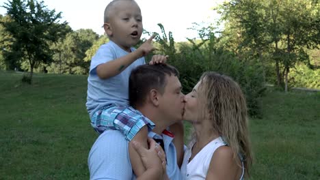 Family-walks-in-the-Park.-The-little-boy-sits-on-his-father's-shoulders,-leans-down-and-kisses-his-mother,-then-shows-his-hand-to-the-side.-Parents-kiss,-look-away-and-smile.