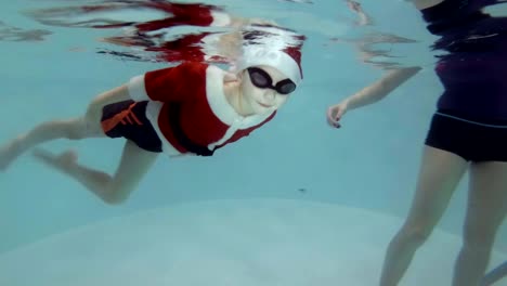 A-little-boy-dressed-as-Santa-Claus-goes-in-for-sports-with-a-coach-in-the-pool.-He-swims-underwater-to-the-stairs-in-swimming-goggles-and-looks-into-the-camera.