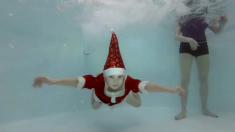 Mamá-ayuda-a-un-niño-pequeño-disfrazado-de-Santa-Claus-para-bucear-bajo-el-agua-en-la-piscina-infantil.-El-niño-nada,-sonríe-y-mira-a-la-cámara.-Cámara-lenta.