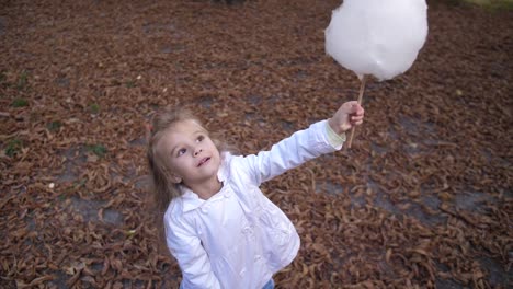 Kleine-blonde-Mädchen-isst-süße-Zuckerwatte-im-Stadtpark.-Schöne-kleine-Mädchen-essen-Zuckerwatte.-Kind-essen-Zuckerwatte-grünen-Wald-auf-den-Migrationshintergrund.-4-k.-Slow-Motion