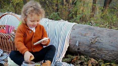 Cute,-curly-haired-toddler-eating-cookies-and-watching-the-phone-in-the-park-on-a-picnic