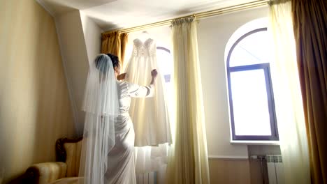 near-window,-in-room,-white-wedding-dress-hanging-on-the-window-eaves.-the-bride,-a-beautiful-girl-in-a-white-peignoir,-a-bathrobe,-is-considering-her-wedding-dress,-preparing-for-the-wedding