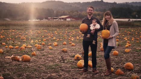 Portrait-of-a-young-family-at-a-pumpkin-patch,-mom-and-dad-holding-pumpkins,-wide-angle-with-lens-flare