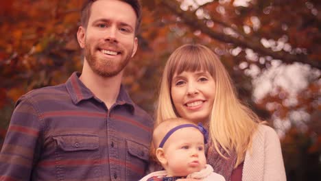 Close-up-portrait-of-a-young-family-standing-under-a-tree-with-colorful-autumn-leaves,-with-beautiful-bokeh