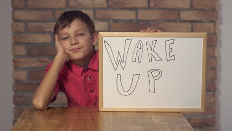 child-sitting-at-the-desk-holding-flipchart-with-lettering-wake-up-on-the-background-red-brick-wall