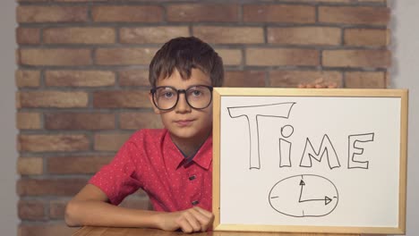 child-sitting-at-the-desk-holding-flipchart-with-lettering-time-on-the-background-red-brick-wall