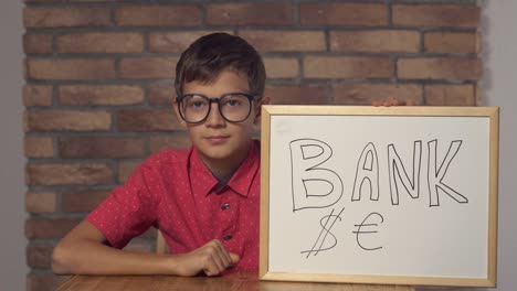 child-sitting-at-the-desk-holding-flipchart-with-lettering-bank-on-the-background-red-brick-wall