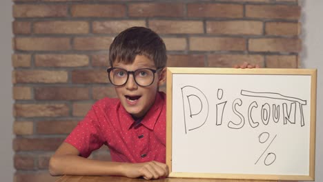 child-sitting-at-the-desk-holding-flipchart-with-lettering-discount-on-the-background-red-brick-wall