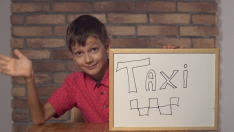 niño-sentado-en-el-escritorio-sosteniendo-rotafolio-con-letras-taxi-en-el-fondo-de-pared-de-ladrillo-rojo