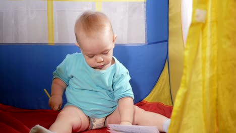 Attractive-baby-playing-in-the-children's-colorful-tent-in-the-house.-The-boy-carefully-draws-in-pencil-notebook