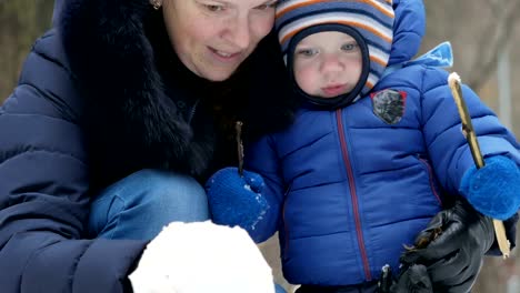 Bebé-y-la-madre-atractiva-esculpe-pequeño-muñeco-de-nieve-en-el-bosque-de-invierno.-Diversión-en-familia-para-las-vacaciones-de-Navidad