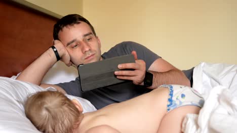 Father-reads-the-news-on-the-tablet-near-the-sleeping-baby.-A-boy-resting-in-the-foreground-of-a-house-on-a-bed