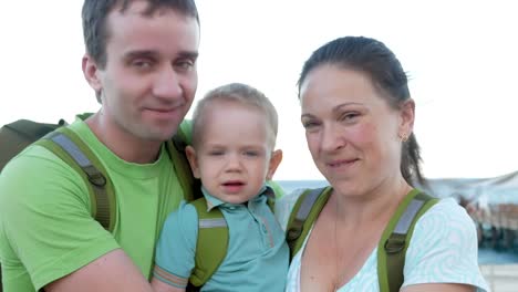 Eine-junge-Familie-mit-einem-Kleinkind-in-die-Kamera-schaut-und-lächelt.-Jeder-Mensch-hat-Rucksäcke-auf-dem-Rücken.-Das-Meer-und-den-Strand-hinter