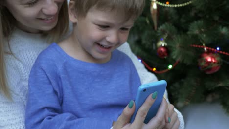 Niño-feliz-y-su-madre-en-el-teléfono-inteligente-cerca-de-árbol-de-Navidad