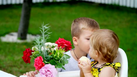 summer,-in-the-garden.-the-four-year-old-boy-gives-a-bouquet-of-flowers-to-his-younger-one-year-old-sister,-brother-kisses-her-sister-on-the-cheek.-The-girl-eats-an-apple