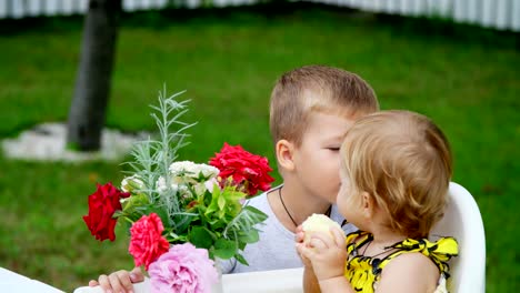 Sommer-im-Garten.-die-vier-jähriger-Junge-gibt-einen-Blumenstrauß-an-seine-jüngeren-ein-Jahr-alte-Schwester,-Bruder-Schwester-auf-die-Wange-küsst.-Das-Mädchen-isst-einen-Apfel