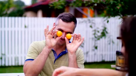 summer,-in-the-garden.-a-man,-father,-having-fun-with-his-family-outdoors,-twisting,-applying-mandarins-to-his-eyes.-family-holidays,-lunch-on-the-nature,-in-the-courtyard