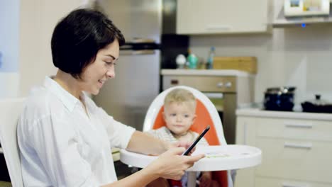 Mutter-nutzt-Telefon-während-kleines-Baby-sitzt-ruhig-im-Kinderstuhl