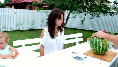 A-young-family-with-two-little-funny-children-sitting-at-a-dinner-table-in-the-garden,-in-summer.-Dad-cuts-a-large-watermelon,-is-going-to-treat-his-family.-lunch-with-the-family