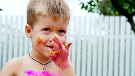 a-little-child,-a-four-year-old-boy-playing,-painting-with-finger-paints,-decorating-himself,-in-the-garden,-sitting-on-a-blanket,-on-grass,-lawn,-in-the-summer.-he's-having-fun