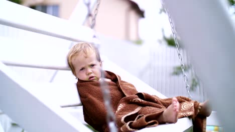 ein-ein-Jahr-alte-Mädchen,-eingewickelt-in-ein-Handtuch-mit-einem-schmutzigen-Gesicht,-einsam-sitzt-auf-einer-Schaukel-im-Garten,-im-Sommer.-Sie-hat-einen-traurigen-Blick.-Sie-will-schlafen