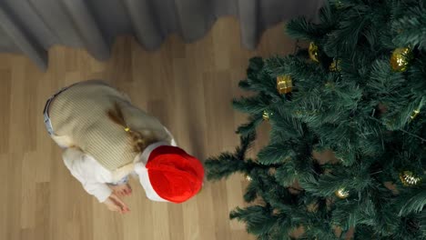 Feliz-joven-de-sombrero-rojo-de-Santa-divertida-y-feliz.-Chica-blanca-decoración-árbol-de-Navidad.-Bola-de-oro-caen-al-piso,-cerca-de-árbol-de-Navidad.-Juguetes-decorativos-de-chica-reparación-esfera-dorada.