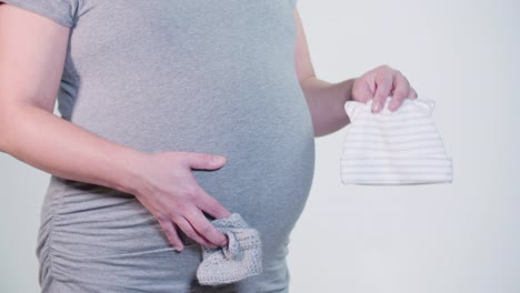 Pregnant-Mother-Holding-Baby-Booties-Hat-in-Hands-Above-Bulging-Belly